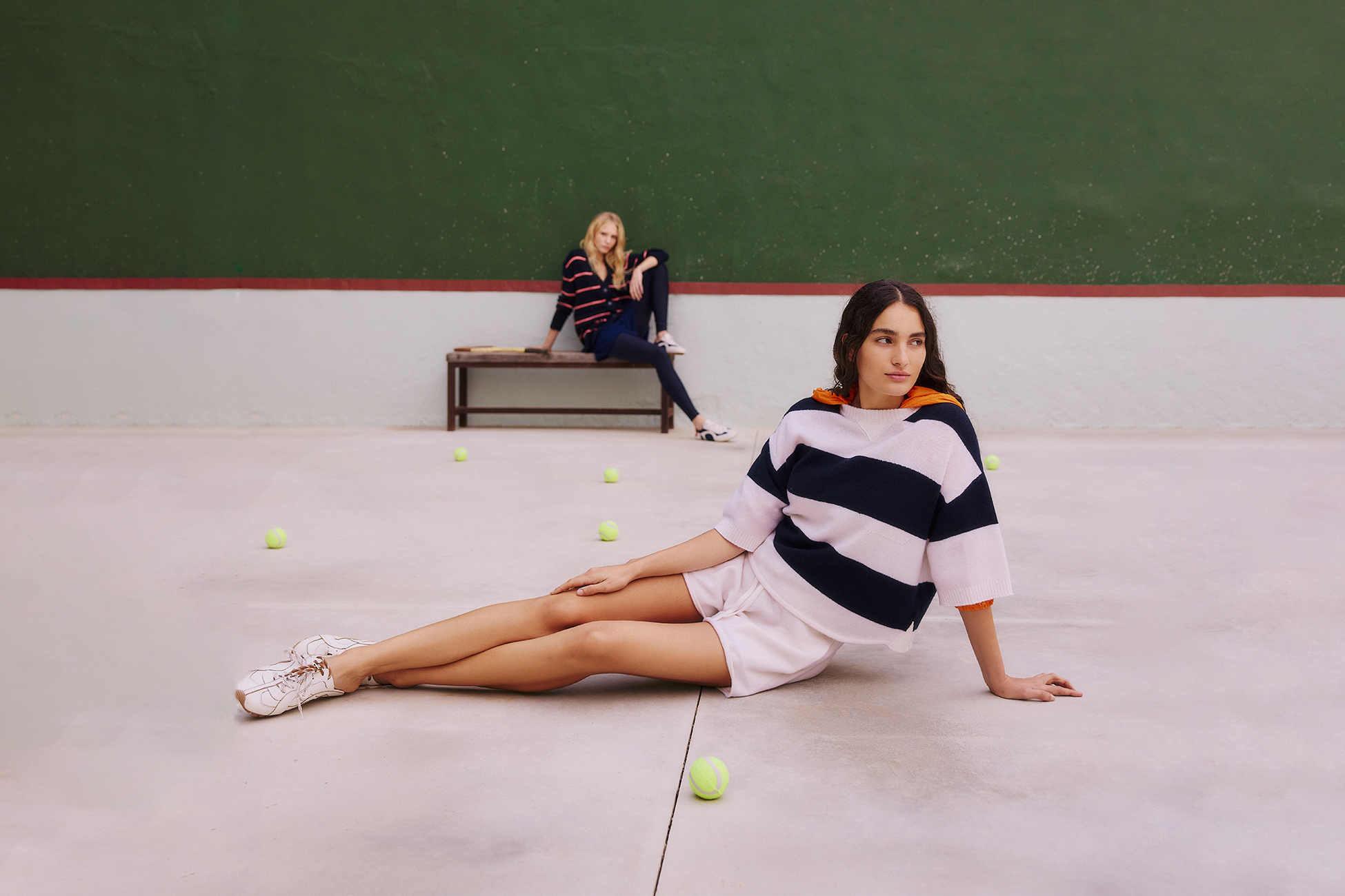 A woman in a navy and white striped sweater and white shorts sits on a light-colored court with tennis balls, while another woman sits on a bench in the background against a green wall.