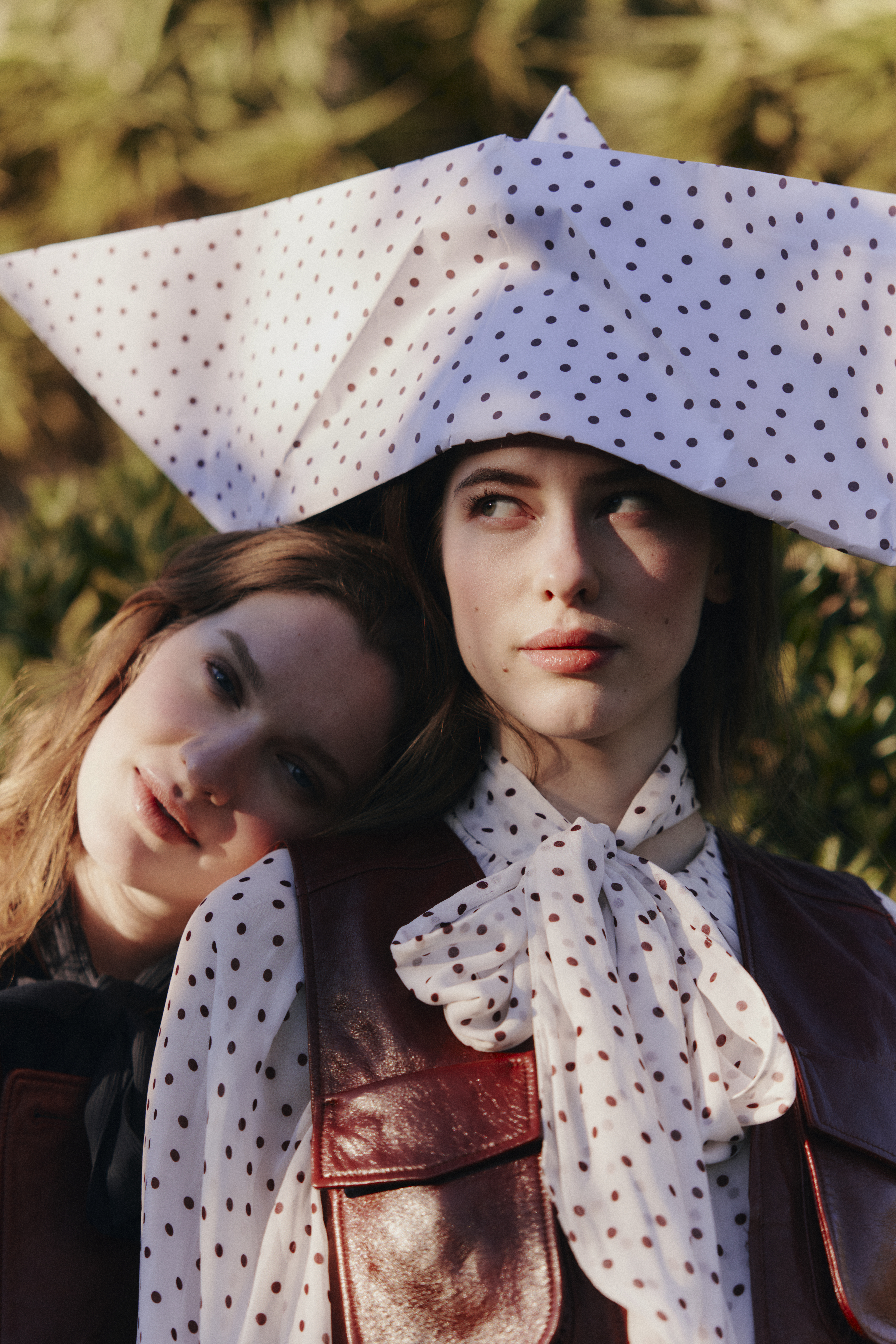 Two Models posing together wearing leather pieces and a blouse with a polka dot print. One of the models is balancing a giant paper boat in the same polka dot print on her head.