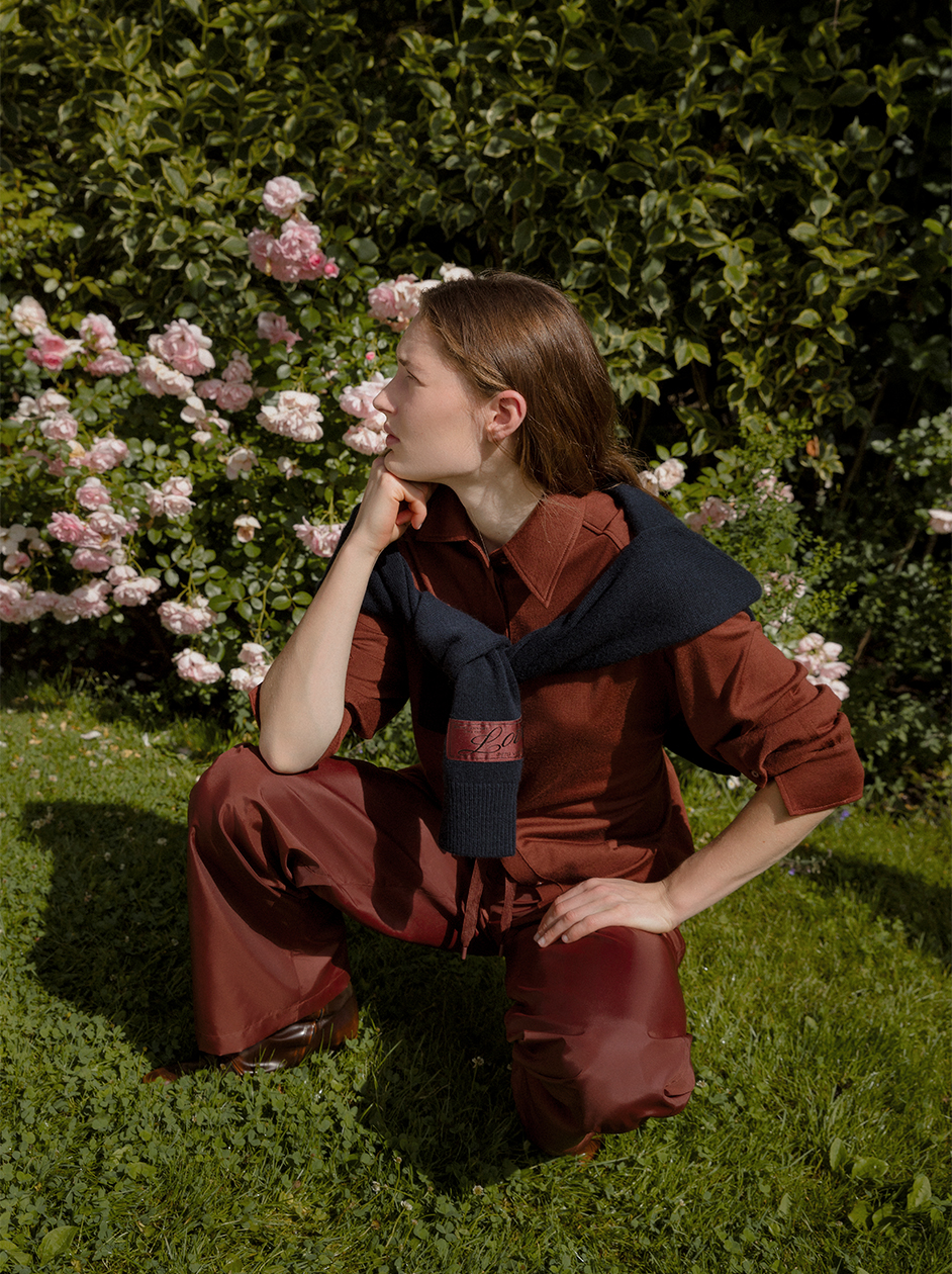 Model in DOROTHEE SCHUMACHER wearing a bordeaux Merino Shirt and Nylon Pants while posing in a garden