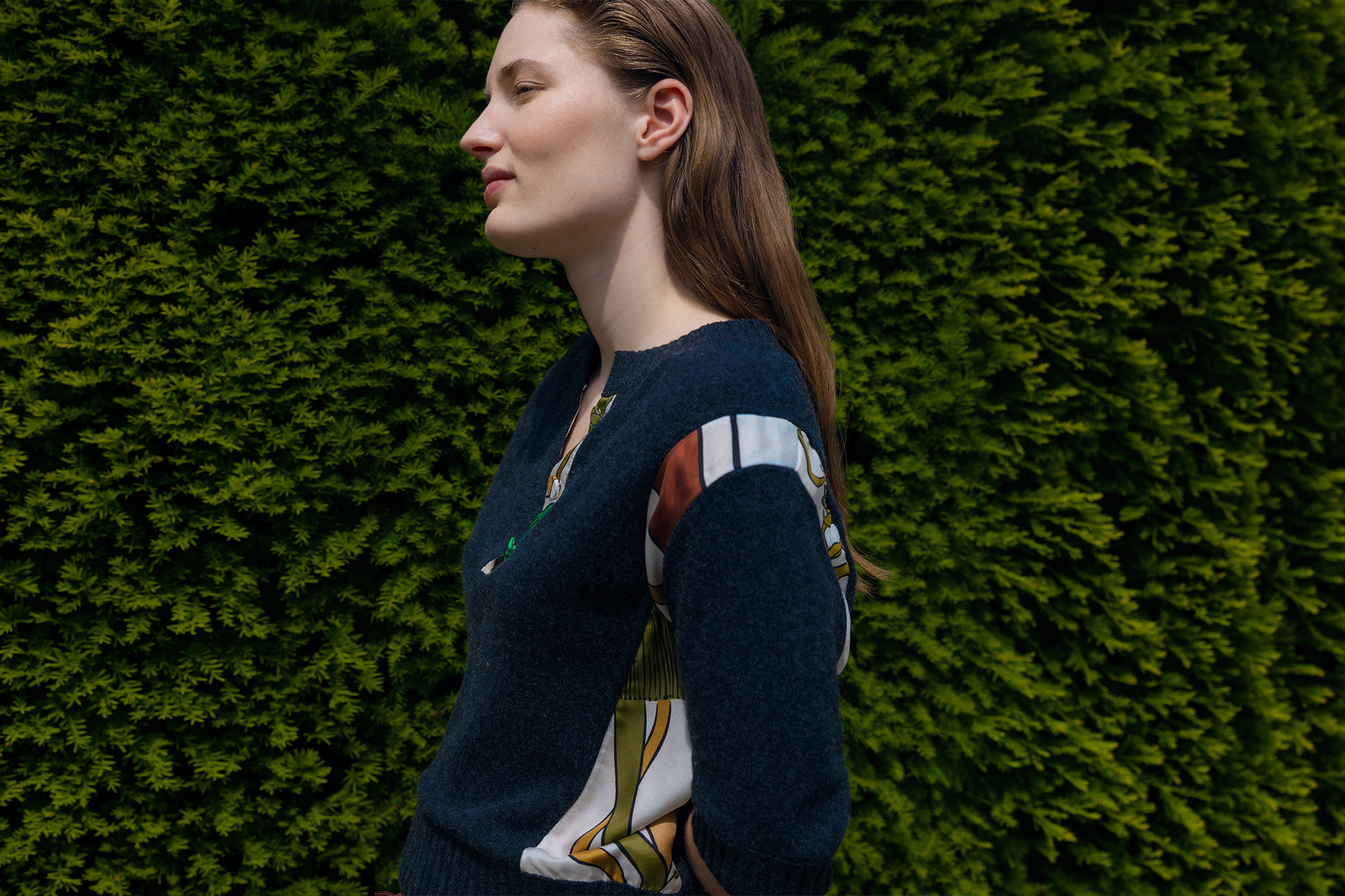 A female Model wearing a Knit Sweater with satin patchwork sleeves from DOROTHEE SCHUMACHER standing in front of a bush