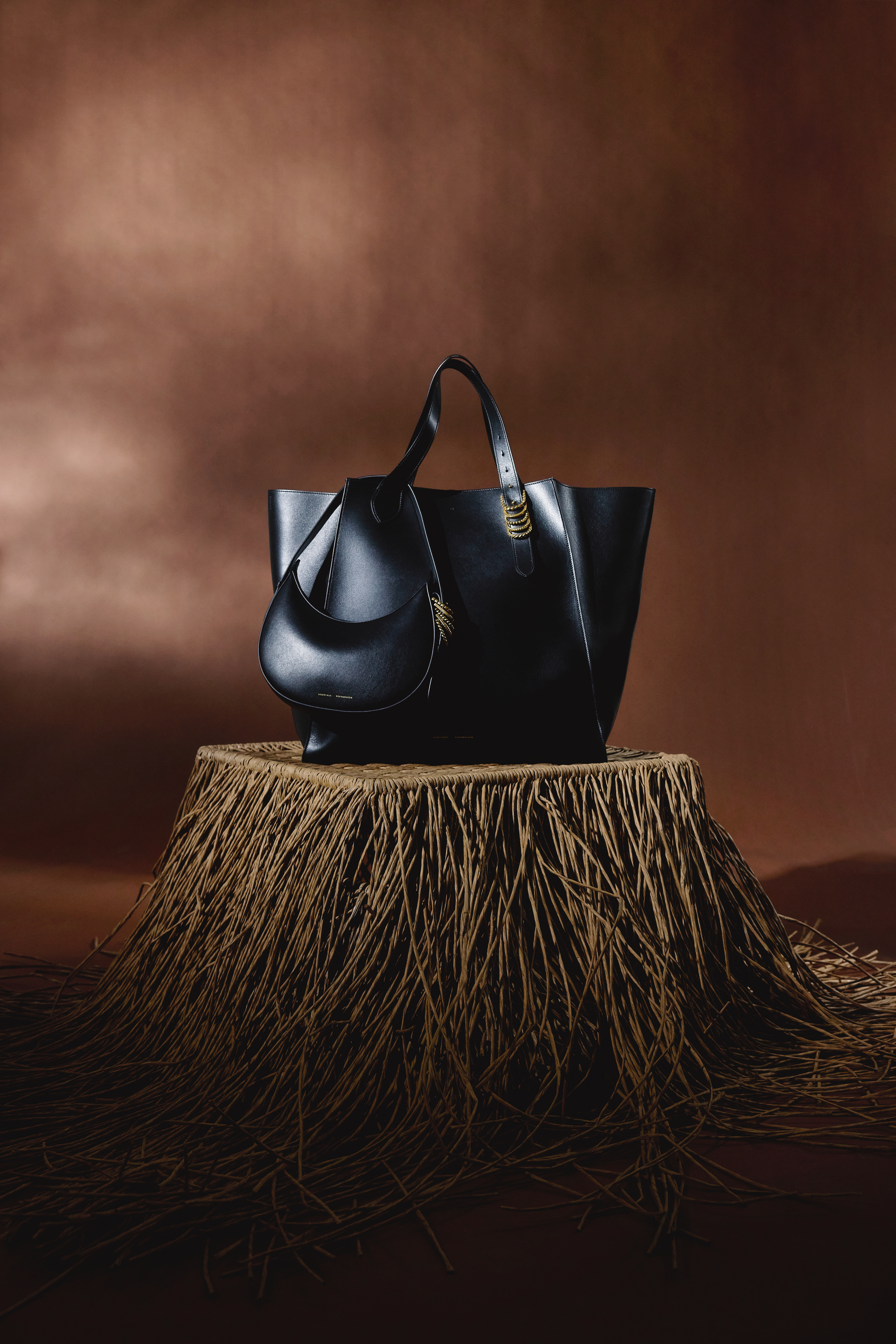 a black half moon leather bag an a black tote bag on a strawy table