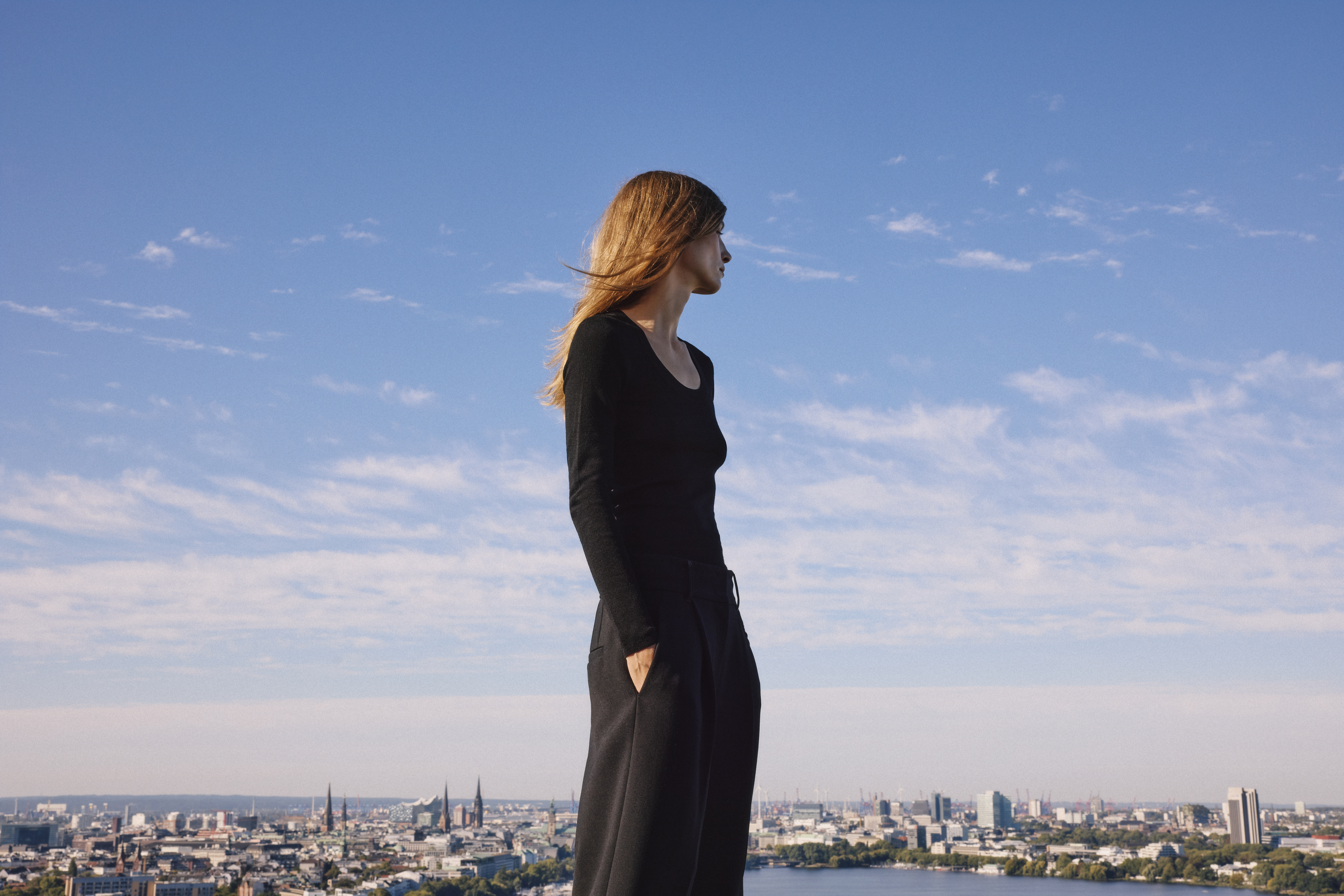 a woman in an all black outfit standing in the front of a city skyline with her hands in the pockets