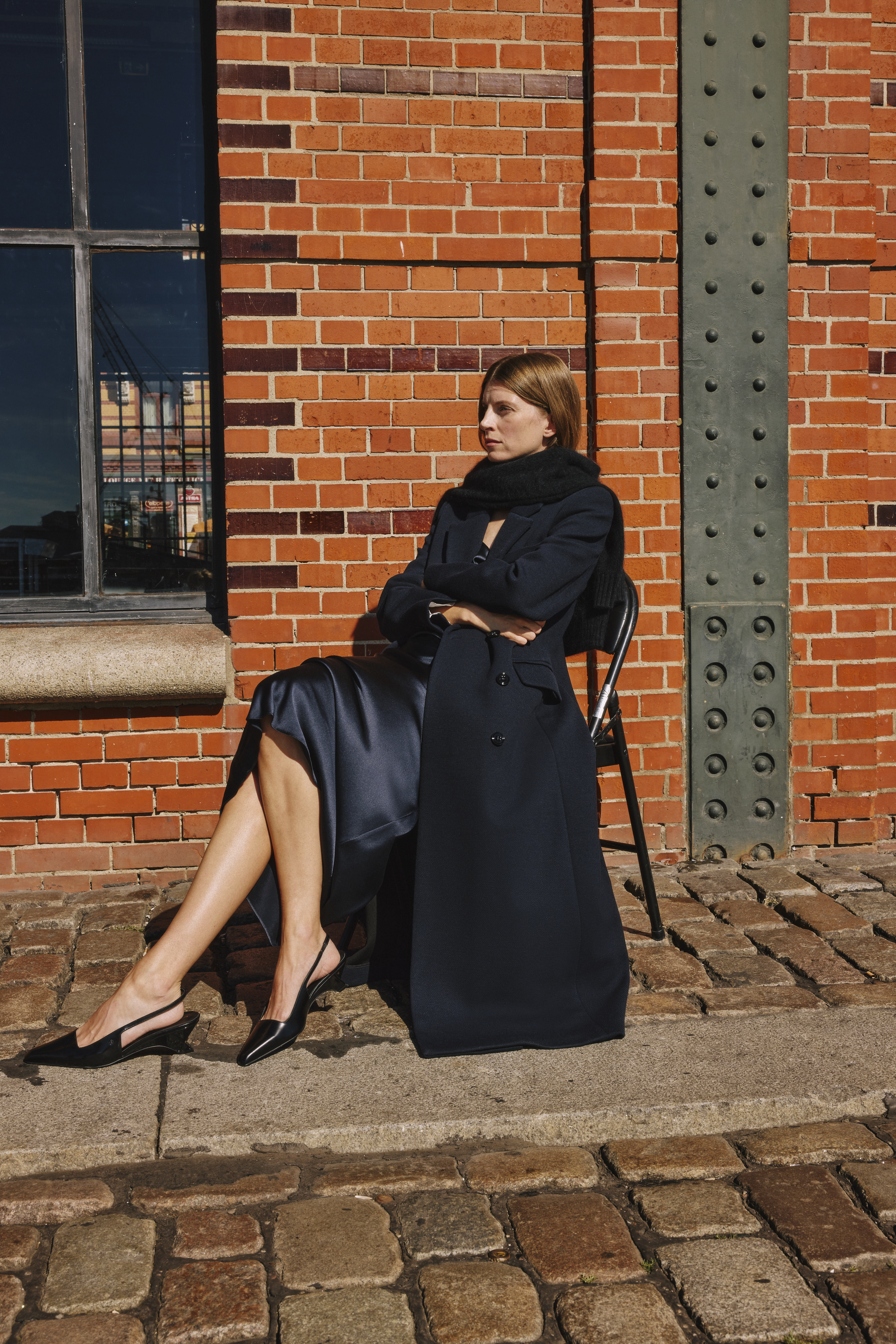 a woman sitting in a blue coat, underneath she´s wearing a blue satin dress and black sling back pumps
