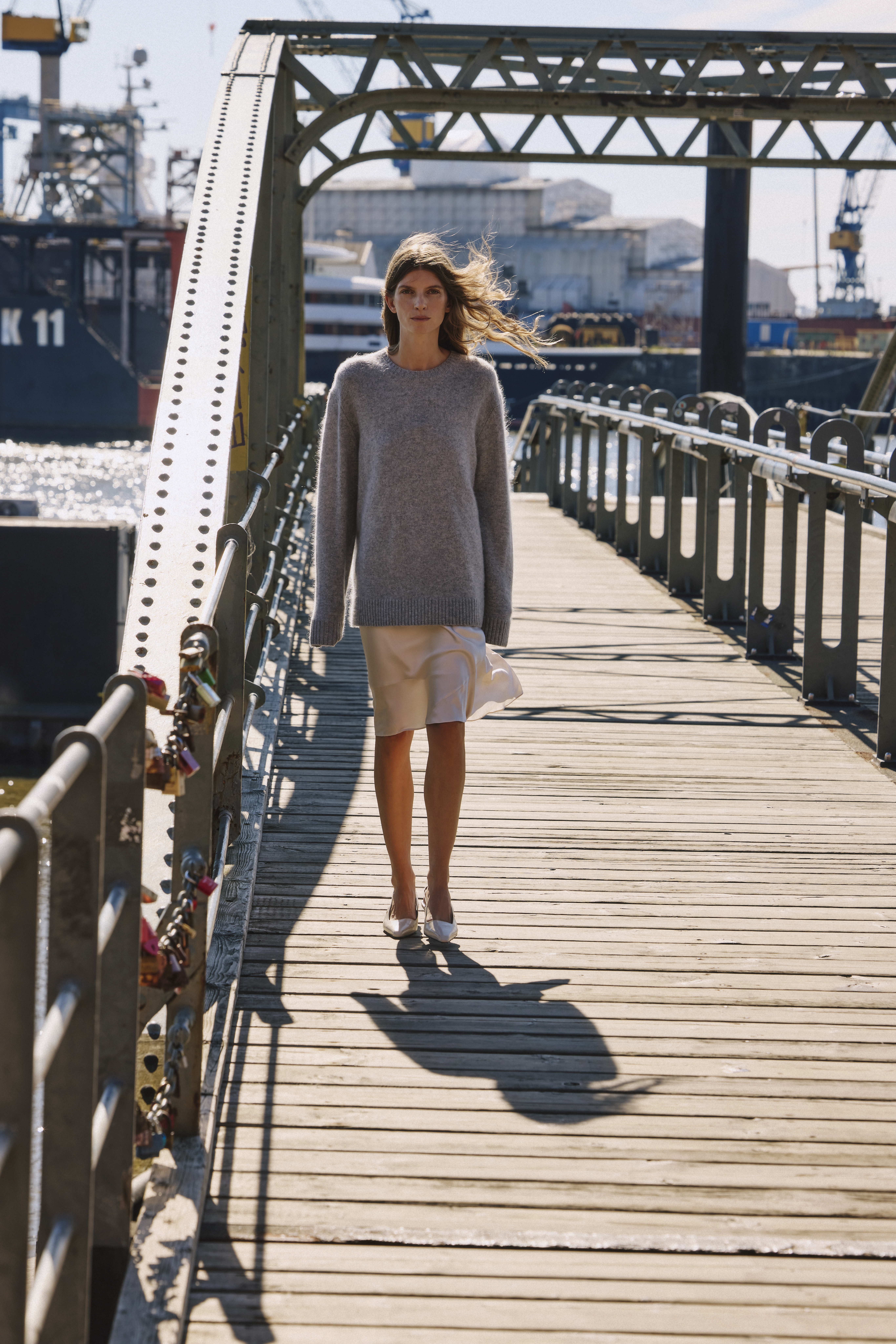 a woman is standing on a bridge wearing a greay pullover, a satin skirt in white and white sling back heels