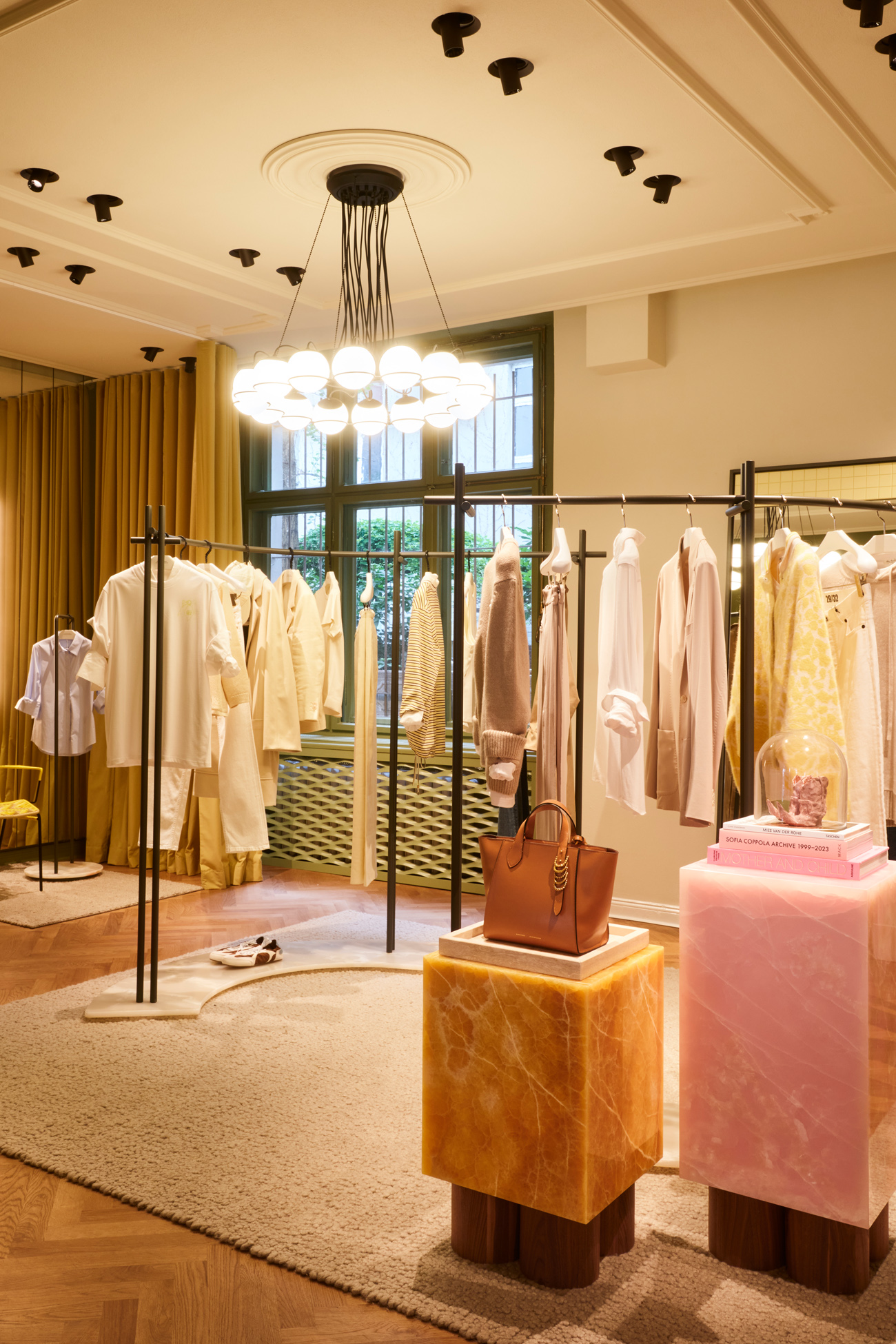 The warm and inviting showroom of the DOROTHEE SCHUMACHER Berlin boutique, showcasing sophisticated ready-to-wear garments on elegant brass racks, artistic marble pedestals, and a modern circular chandelier.