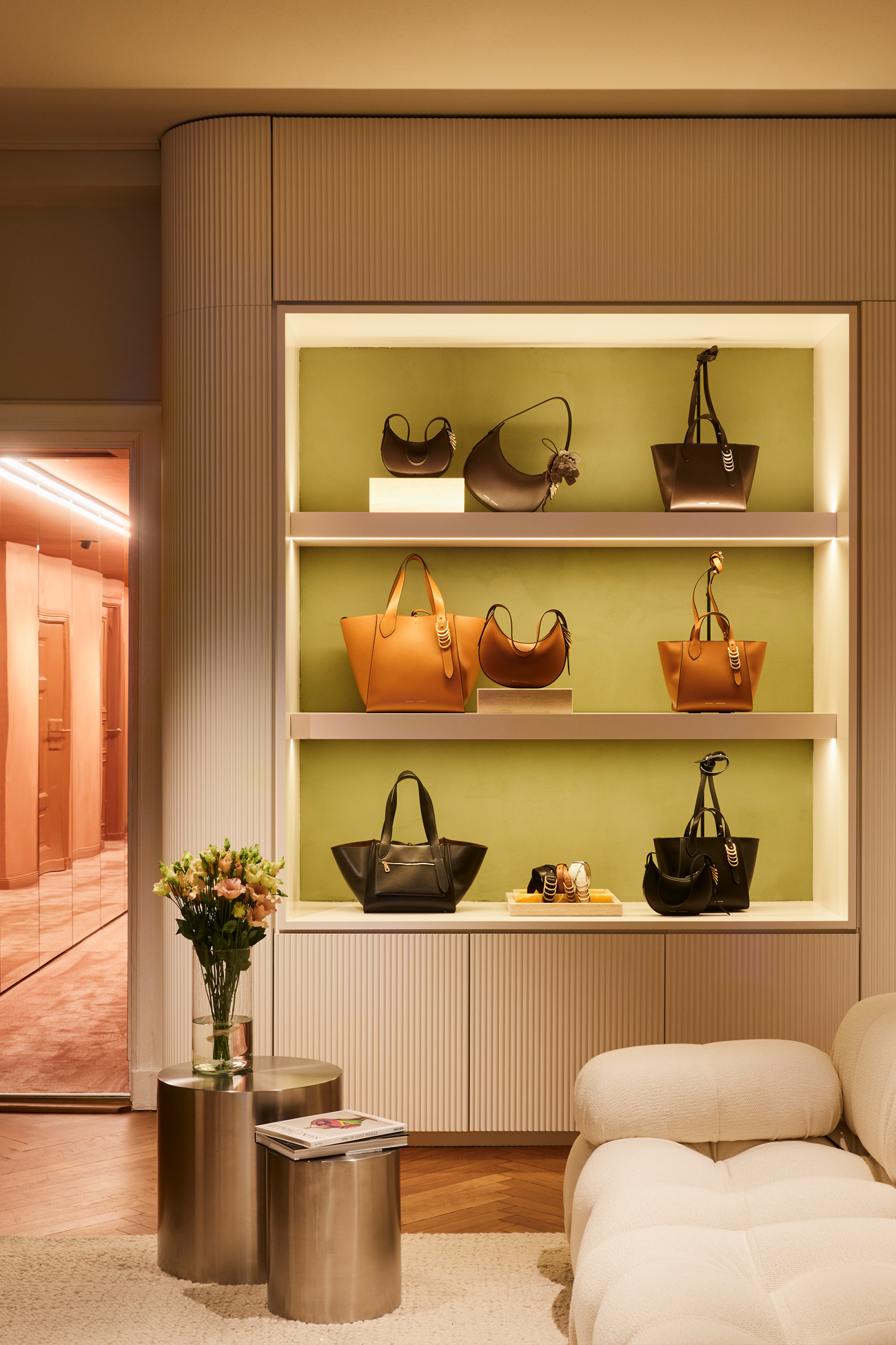 A luxurious lounge and display area in the DOROTHEE SCHUMACHER Berlin store, featuring illuminated built-in shelves showcasing premium leather handbags, a modern white sofa, and an elegant mirror reflection.