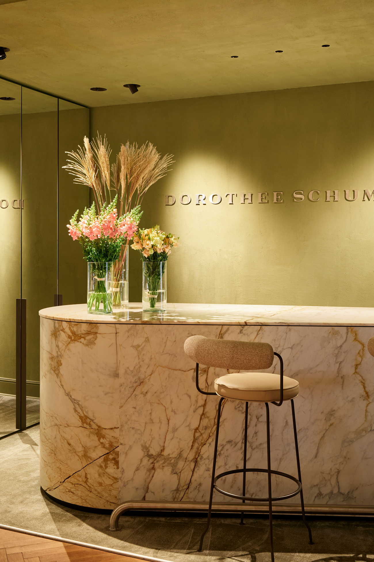 An elegant and sophisticated marble reception counter inside the DOROTHEE SCHUMACHER Berlin store, adorned with vibrant floral arrangements and an embossed golden logo on a refined olive-green plaster wall.
