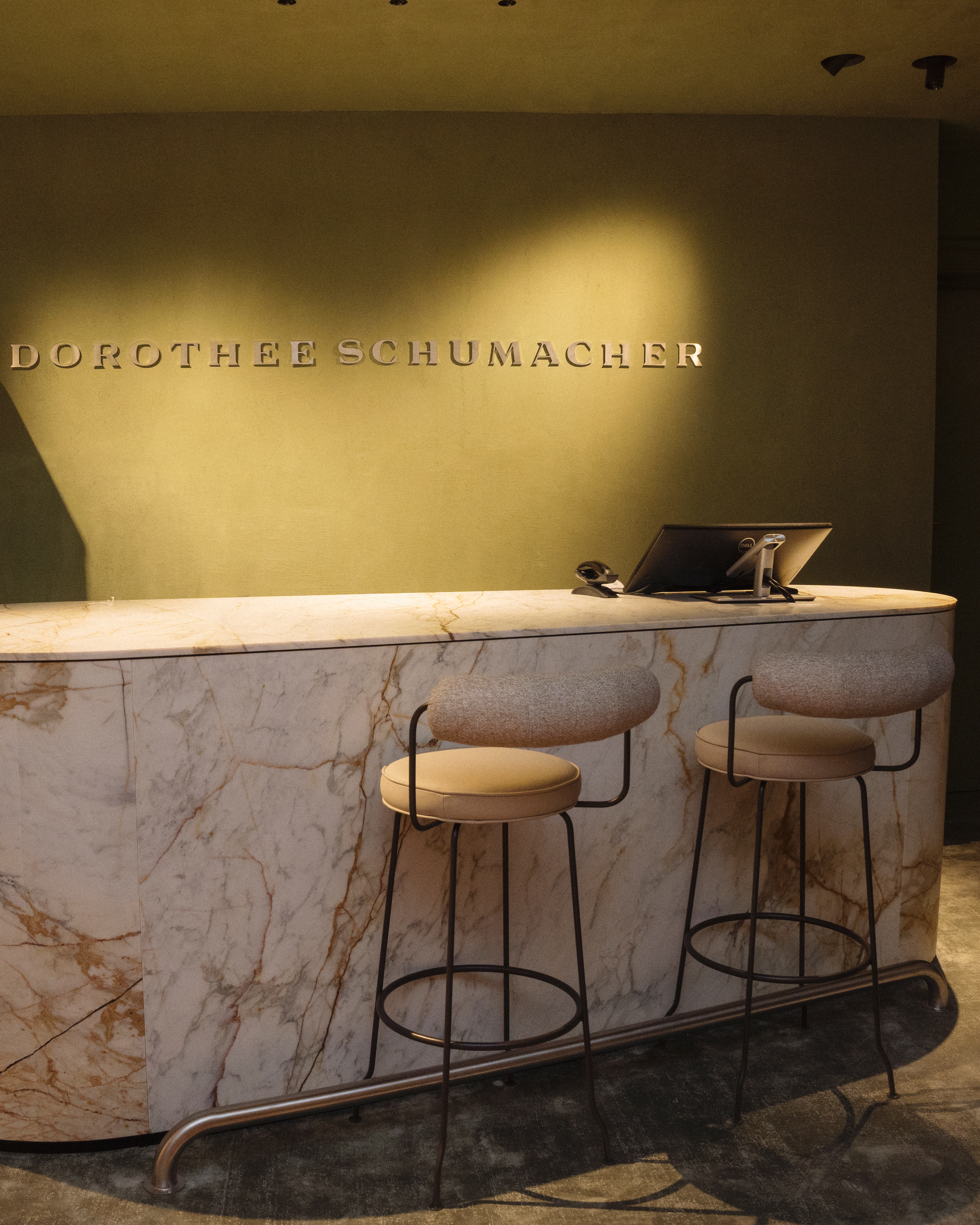 Marmor-Empfangstheke im Dorothee Schumacher Geschäft mit dem beleuchteten 'DOROTHEE SCHUMACHER' Logo an der Wand und zwei Barhockern.