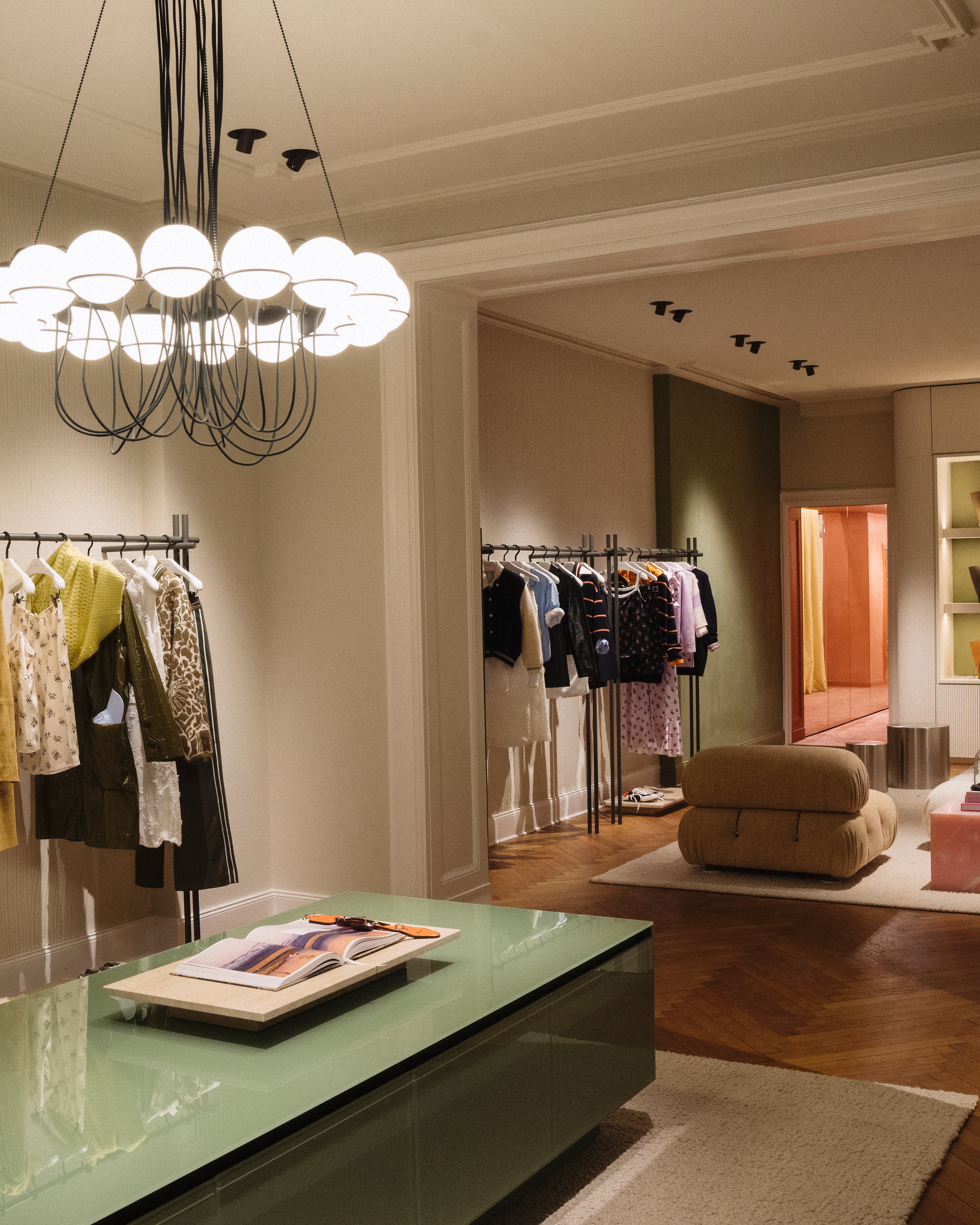 Helles Interieur des Dorothee Schumacher Geschäfts mit einem auffälligen Kronleuchter, Kleiderständern voller Damenmode, einem grünen Tisch mit einem Magazin und einer gemütlichen Sitzecke.