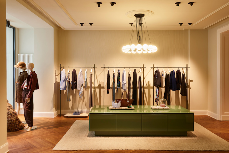 The warm and inviting showroom of the DOROTHEE SCHUMACHER Berlin boutique, showcasing sophisticated ready-to-wear garments on elegant brass racks, artistic marble pedestals, and a modern circular chandelier.
