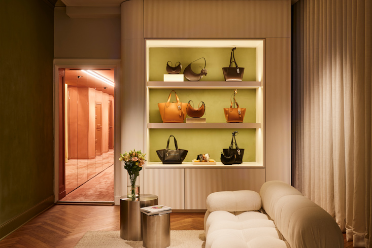 A luxurious lounge and display area in the DOROTHEE SCHUMACHER Berlin store, featuring illuminated built-in shelves showcasing premium leather handbags, a modern white sofa, and an elegant mirror reflection.