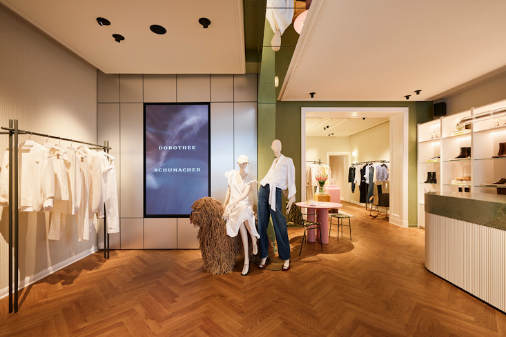 A luxurious lounge and display area in the DOROTHEE SCHUMACHER Berlin store, featuring illuminated built-in shelves showcasing premium leather handbags, a modern white sofa, and an elegant mirror reflection.