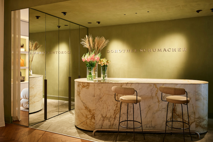 An elegant and sophisticated marble reception counter inside the DOROTHEE SCHUMACHER Berlin store, adorned with vibrant floral arrangements and an embossed golden logo on a refined olive-green plaster wall.