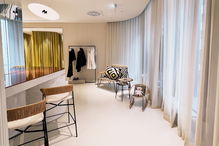 Gemütlicher Lounge-Bereich im Dorothee Schumacher Geschäft in Dresden mit einer Sitzbank, einem Stuhl, einem Kleiderständer und hellen Vorhängen.