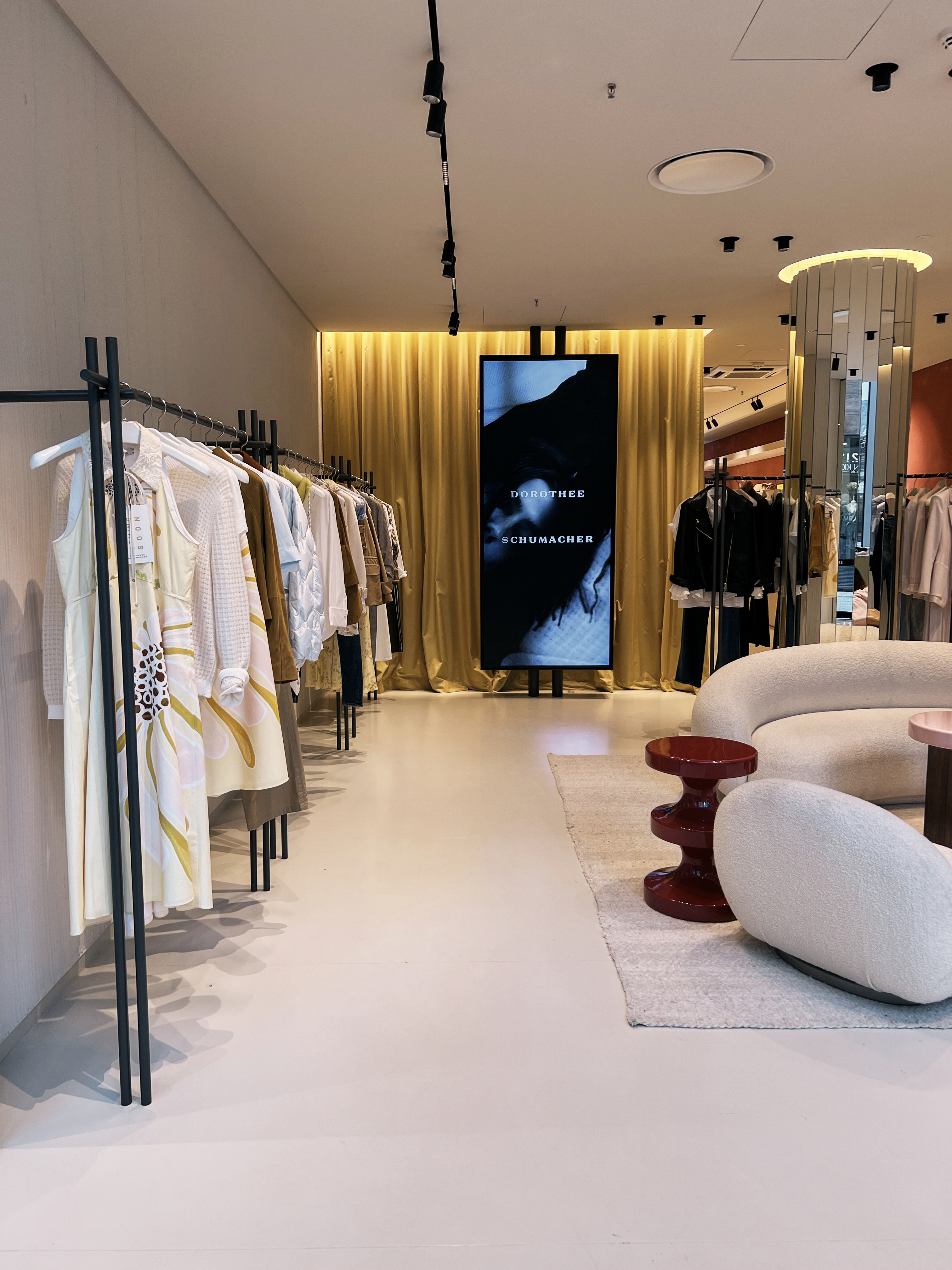 Interior view of a Dorothee Schumacher store featuring elegant clothing racks, a large branded video screen, and cozy boucle furniture.