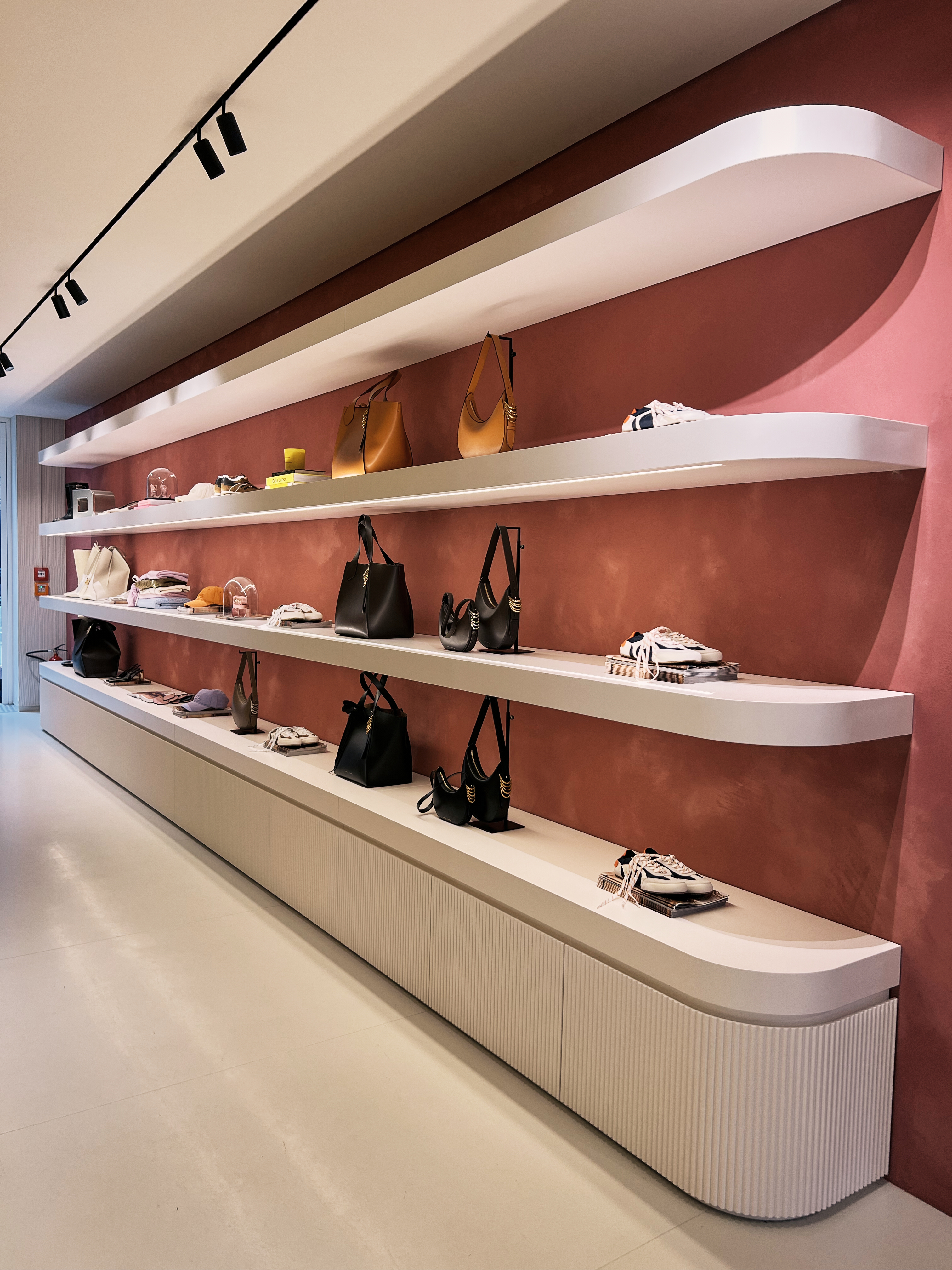 Modern curved wall shelves in a Dorothee Schumacher store displaying a curated selection of handbags and sneakers against a terracotta-colored wall.