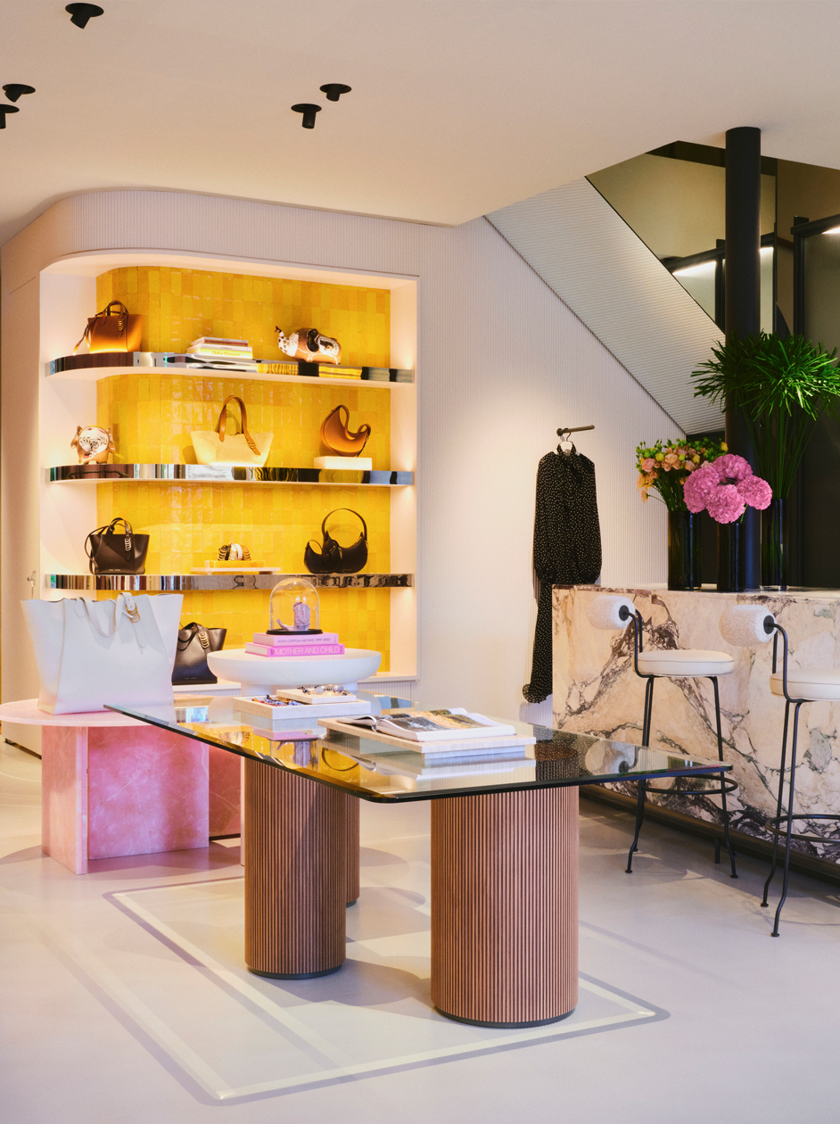 The elegant main display area of the DOROTHEE SCHUMACHER boutique in Munich, featuring a modern glass-top table with fluted wooden columns, a marble service counter with barstools, and a vibrant yellow-backlit wall showcasing luxury handbags.
