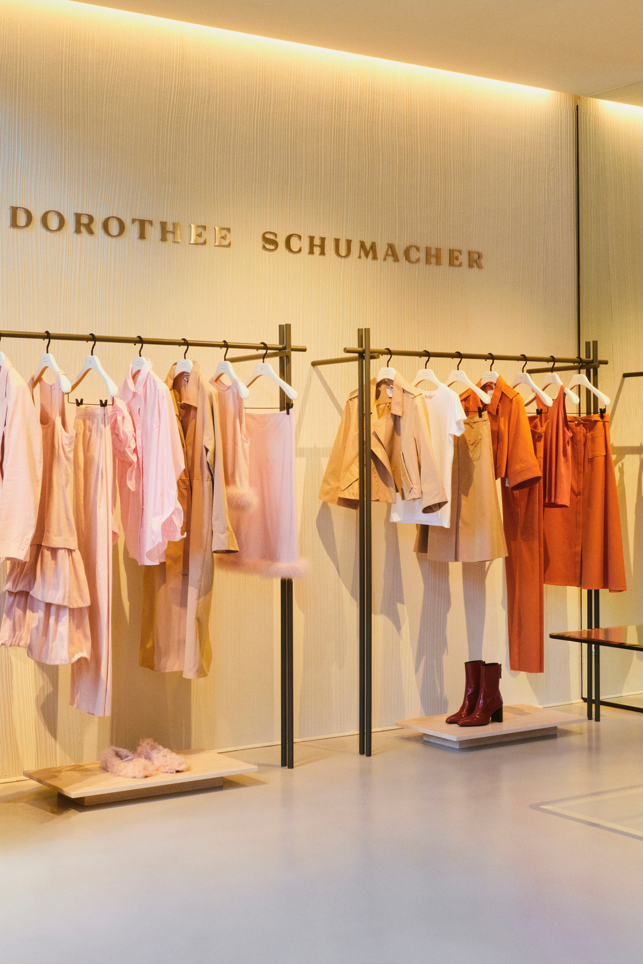 An interior view of the DOROTHEE SCHUMACHER Munich boutique showcasing chic clothing racks under the golden brand wall logo. The racks display premium seasonal garments in warm shades of rose, peach, and terracotta.