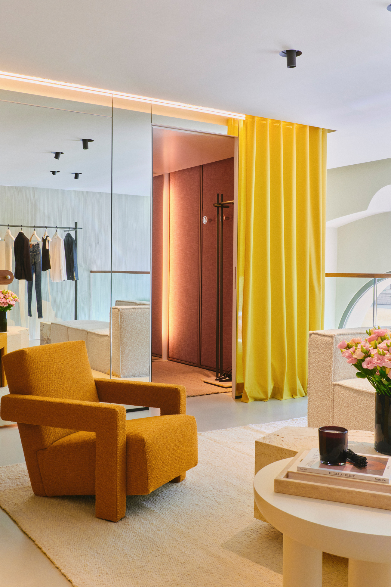 A perspective of the bright main retail floor in the DOROTHEE SCHUMACHER Munich store, highlighting minimalist garment rails, a textured terracotta-colored feature wall, and a central glass-top styling tables.