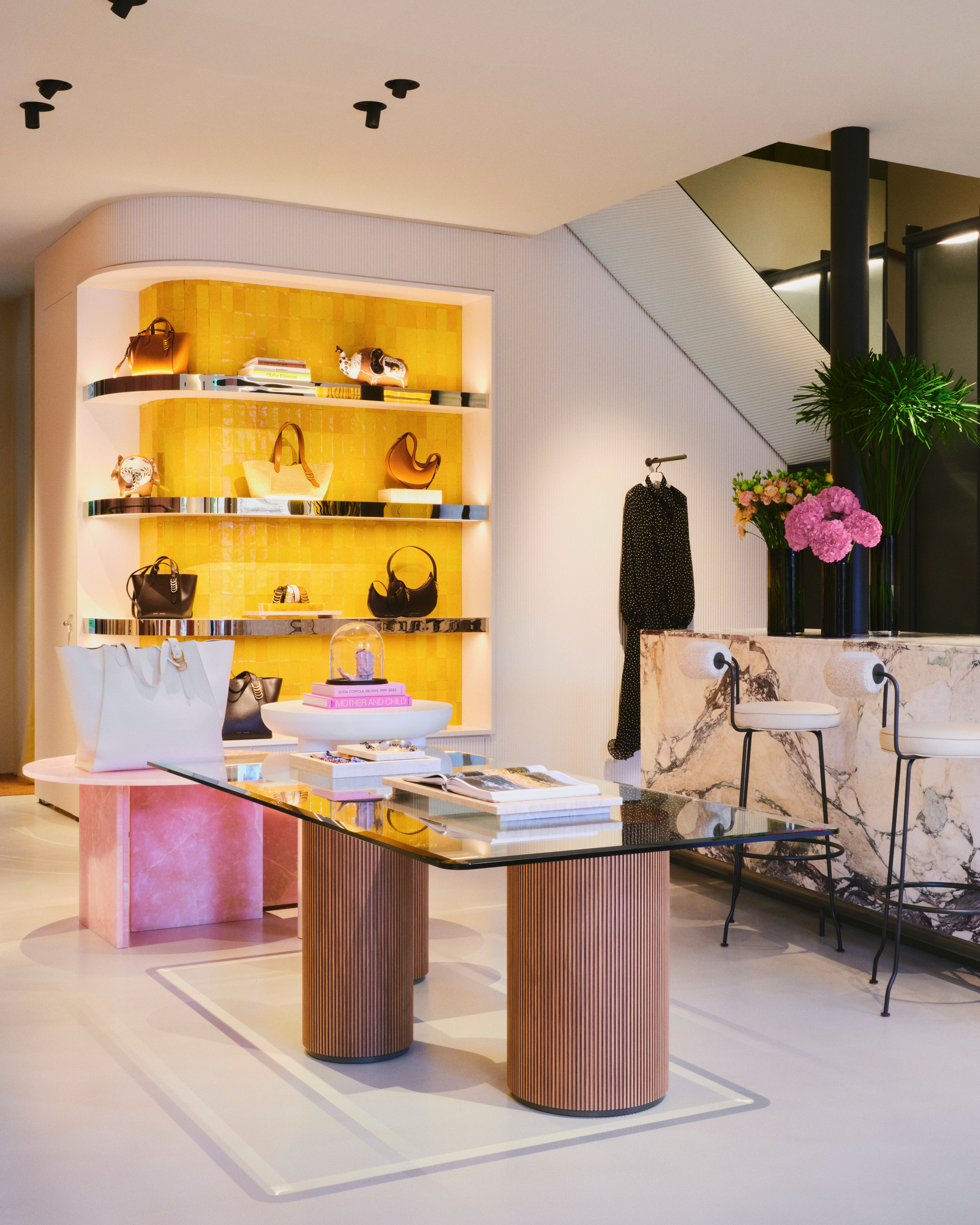 The elegant main display area of the DOROTHEE SCHUMACHER boutique in Munich, featuring a modern glass-top table with fluted wooden columns, a marble service counter with barstools, and a vibrant yellow-backlit wall showcasing luxury handbags.