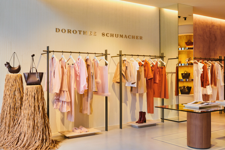An interior view of the DOROTHEE SCHUMACHER Munich boutique showcasing chic clothing racks under the golden brand wall logo. The racks display premium seasonal garments in warm shades of rose, peach, and terracotta.