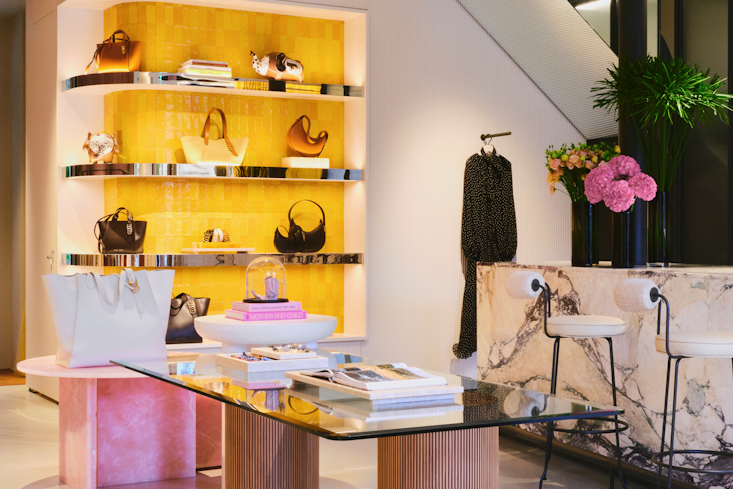 The elegant main display area of the DOROTHEE SCHUMACHER boutique in Munich, featuring a modern glass-top table with fluted wooden columns, a marble service counter with barstools, and a vibrant yellow-backlit wall showcasing luxury handbags."