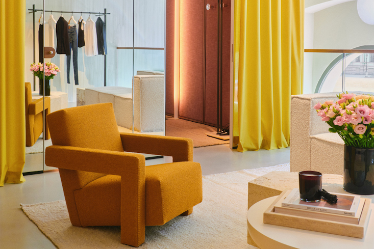 A sophisticated lounge and consultation area inside the DOROTHEE SCHUMACHER Munich store, featuring a plush mustard-yellow armchair, a low round coffee table with fresh flowers, and dramatic, vibrant yellow drapes.