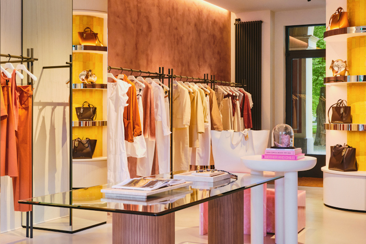 A perspective of the bright main retail floor in the DOROTHEE SCHUMACHER Munich store, highlighting minimalist garment rails, a textured terracotta-colored feature wall, and a central glass-top styling tables.